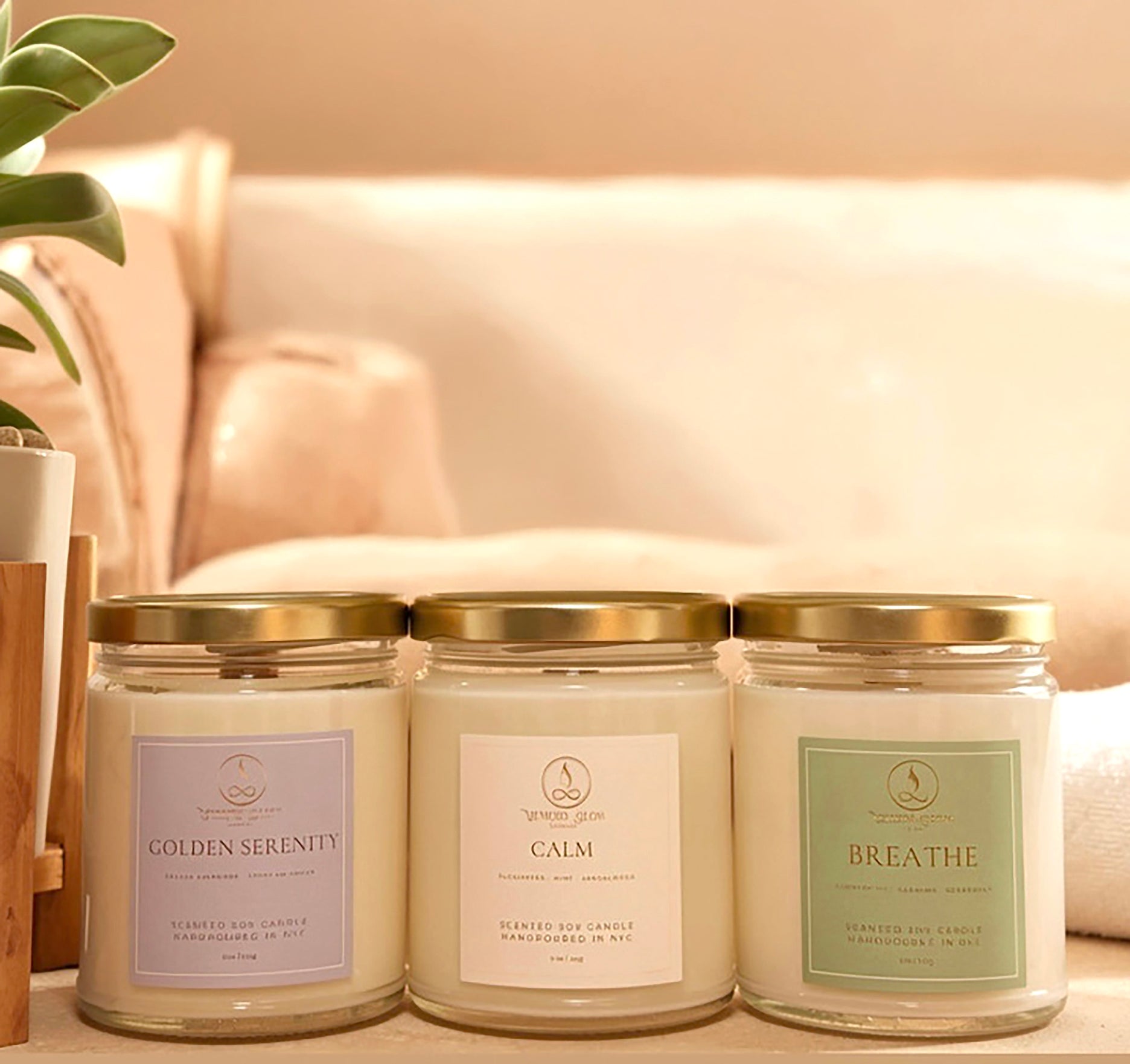 Three scented soy candles in clear glass jars with gold lids are lined up on a wooden surface. The labels read Golden Serenity, Calm, and Breathe, with soft pastel lavender, cream, and sage green designs.