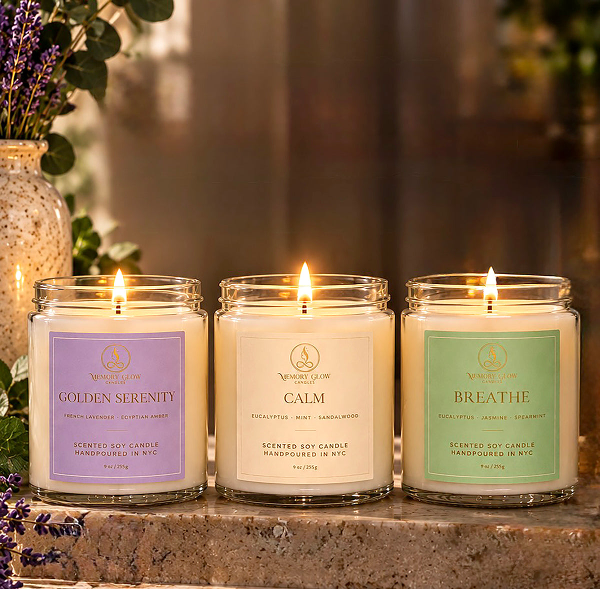 Three lit scented soy candles in clear glass jars are arranged on a stone surface in a warm, softly blurred indoor setting. The candles have lavender, cream, and mint labels reading Golden Serenity, Calm, and Breathe, with the brand name, Memory Glow visible on each label.