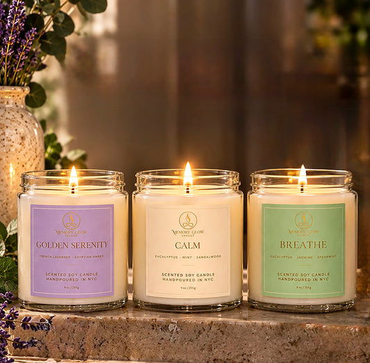 Three lit scented soy candles in clear glass jars are arranged on a stone surface in a warm, softly blurred indoor setting. The candles have lavender, cream, and mint labels reading Golden Serenity, Calm, and Breathe, with the brand name, Memory Glow visible on each label.