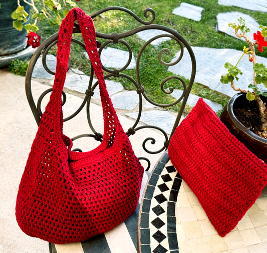 A red crocheted shoulder bag with strap sits on a black wrought-iron cafe chair next to a matching red crocheted zipper pouch, set on a tiled table with a potted plant and green grass in the background.