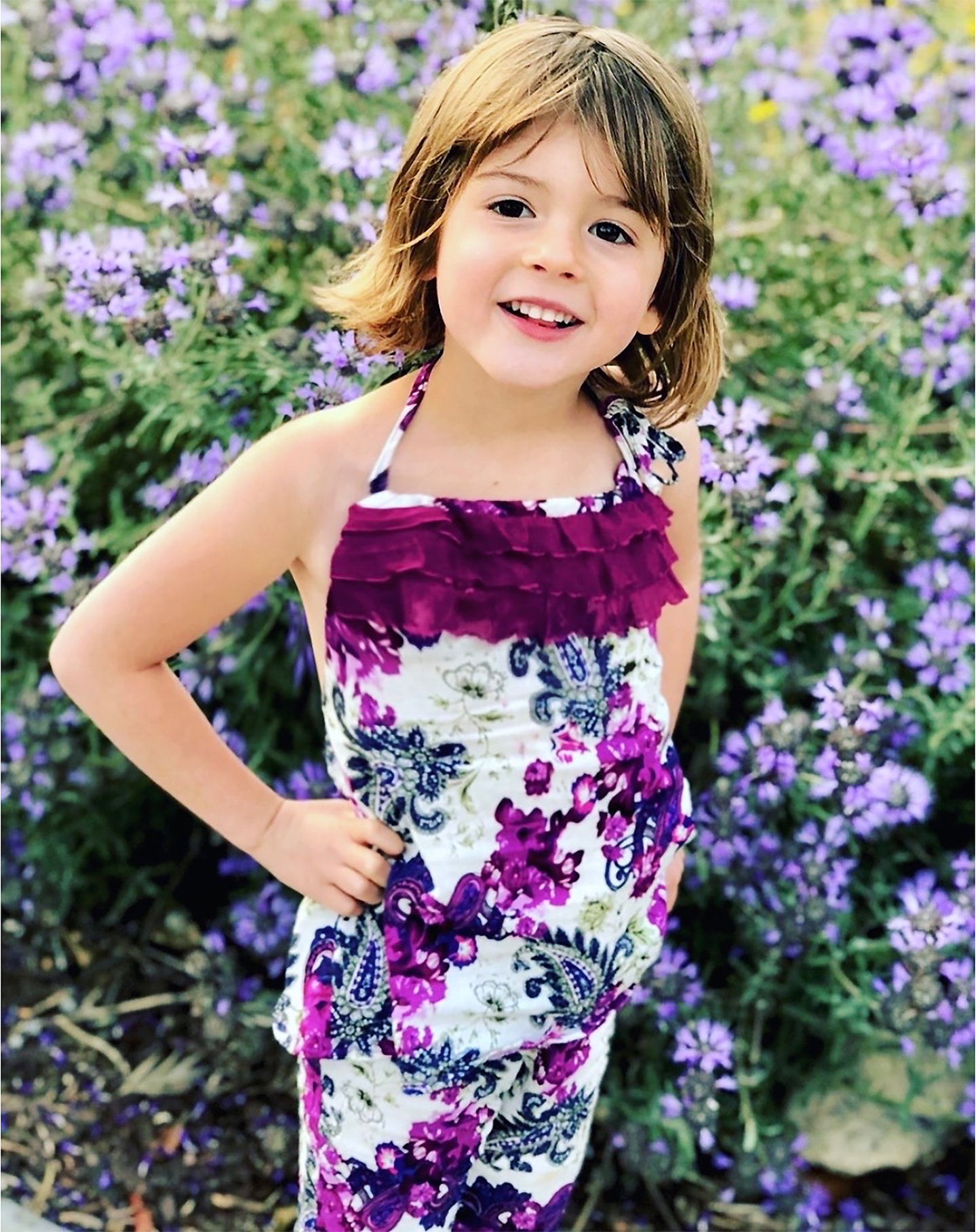 Young girl in a white, purple, blue floral print halter top/ pant set, standing among purple flowers. Best Selling colorful purple paisley-floral print on soft, light weight, cotton woven fabric. A-line halter top with delicate ruffle trim, tie back, and soft elastic around the back to stay in place. Pair with the Gatsby Girl Capri pant for a comfy, flower fun look. 100% cotton woven Hand wash or machine wash delicate cycle, cold. Line dry. Sustainably made in California